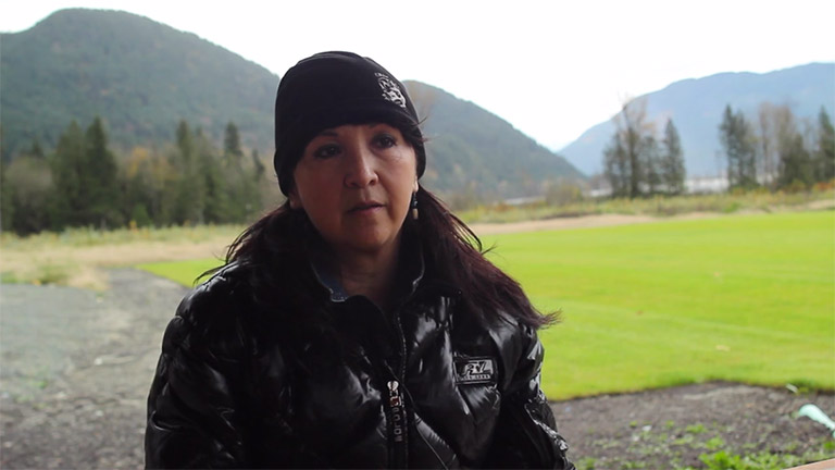 A woman is sitting outside in the center of the image. She is wearing a black coat and toque. In the background there are trees, with large mountains behind.