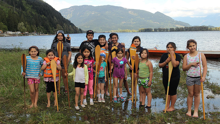 Fourteen young children stand near the shore of a river, smiling, and holding onto canoe paddles that are positioned in front of them vertically. A canoe is behind them. In the background is the river, and green mountains are in the distance.