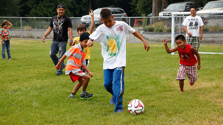Un groupe d’hommes et d’enfants jouent au soccer sur un terrain gazonné. Les enfants courent après l’homme qui dribble le ballon.