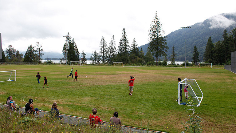 A game of soccer is being played on an outdoor grass field, with trees and mountains in the background. In the foreground, several spectators are leaning against a fence watching the game.