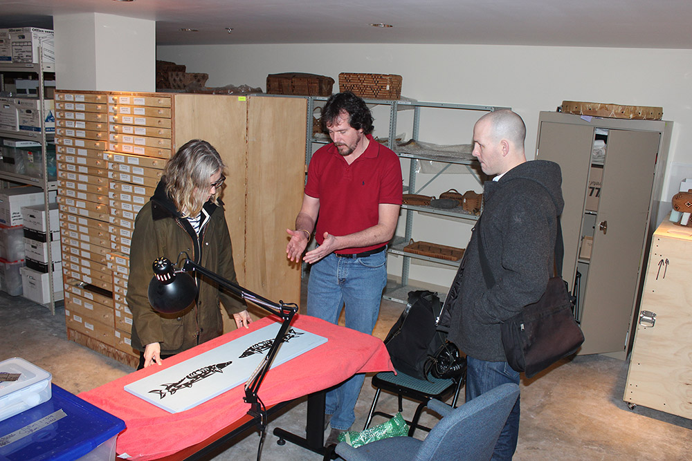 Three people are standing around a table, looking at a painting that is laying on top of an orange cloth. The painting is white, with two black sturgeon fish facing each other.