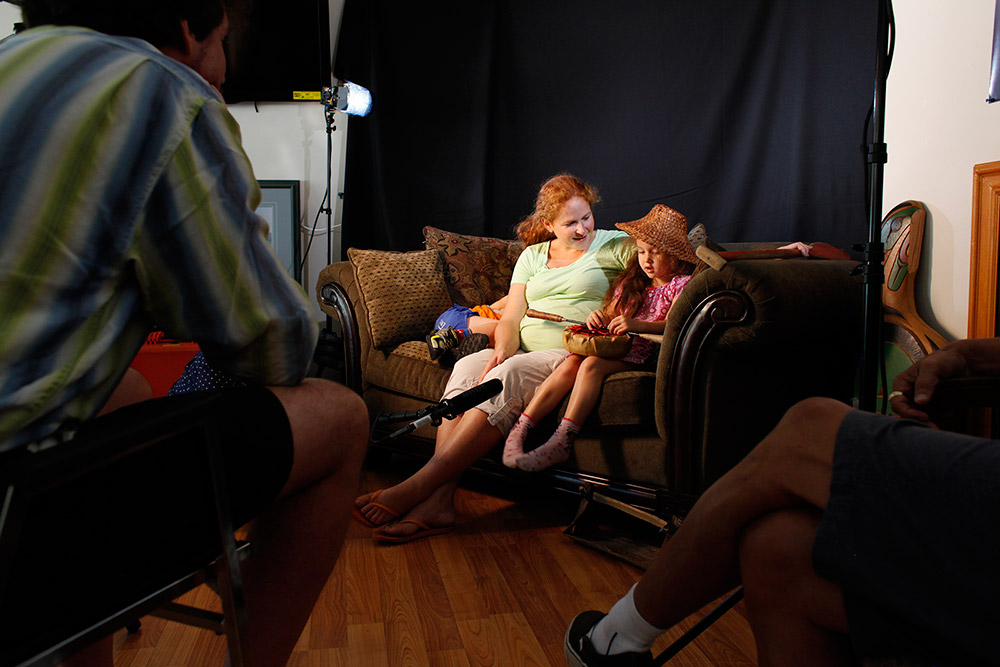 Une femme et une fillette assises sur un divan. Au premier plan, plusieurs personnes sont assises et les regardent.