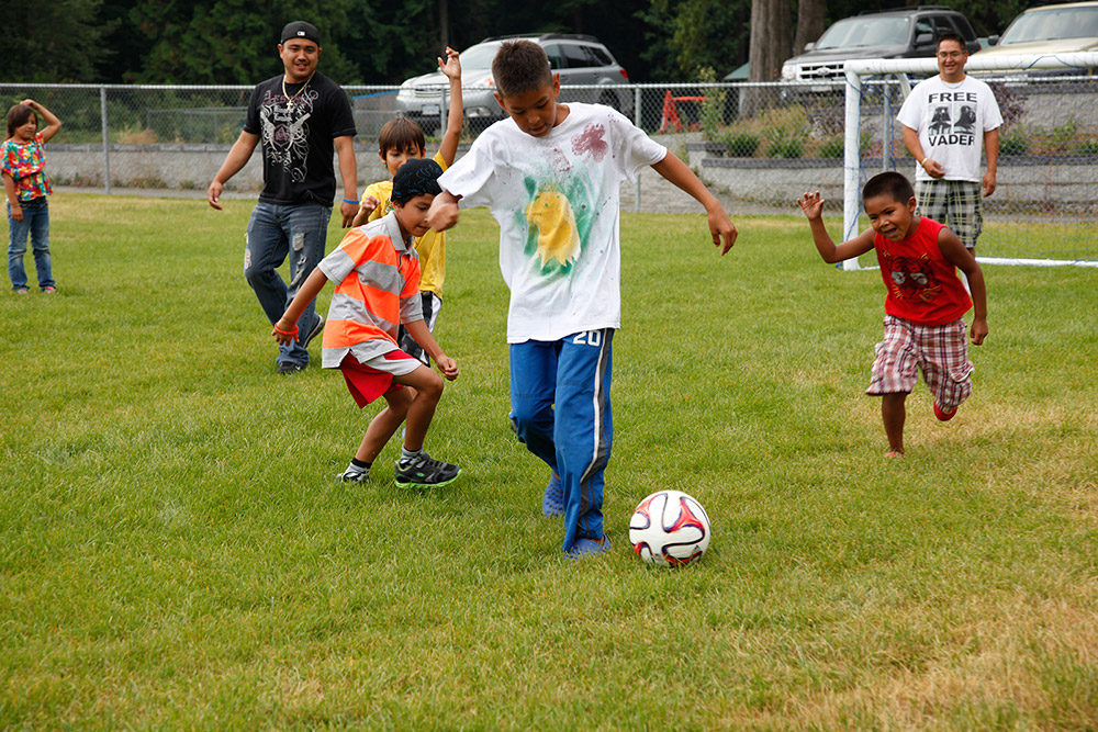 Un groupe d’hommes et d’enfants jouent au soccer sur un terrain gazonné. Les enfants courent après l’homme qui dribble le ballon.