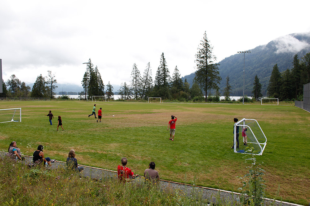 A game of soccer is being played on an outdoor grass field, with trees and mountains in the background. In the foreground, several spectators are leaning against a fence watching the game.