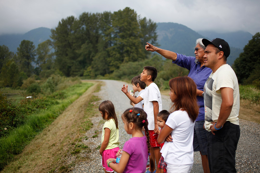 A group of children are gathered on a gravel road, with two men standing behind them. The older man is pointing off in the distance, and the children are looking in that direction.