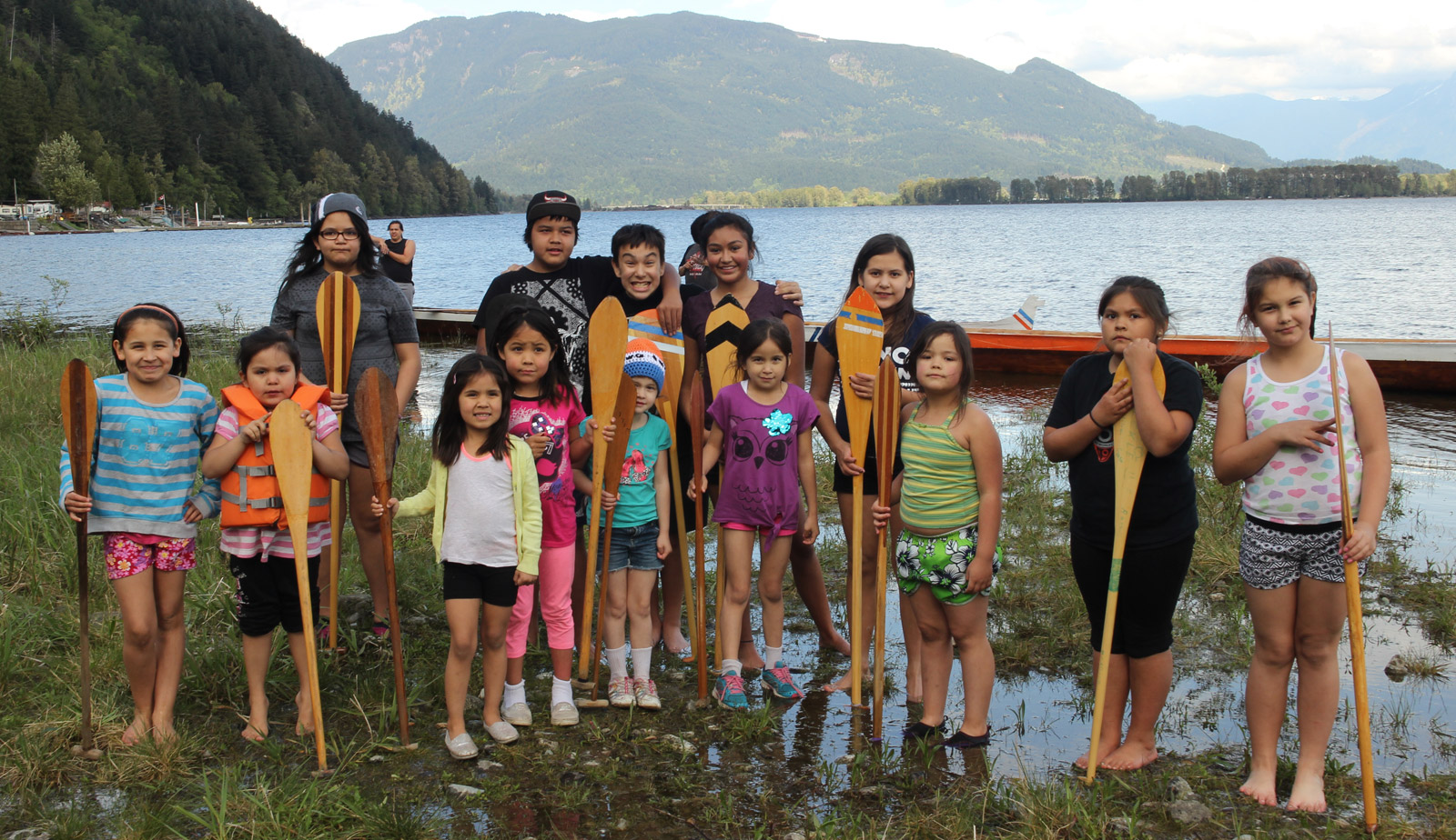 Fourteen young children stand near the shore of a river, smiling, and holding onto canoe paddles that are positioned in front of them vertically. A canoe is behind them. In the background is the river, and green mountains are in the distance. 