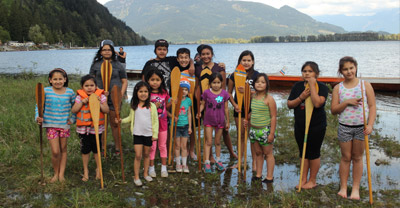 Fourteen young children standing near the shore of a river, smiling, and holding onto canoe paddles that are positioned in front of them vertically. A canoe is behind them. In the background is the river, and green mountains in the distance.