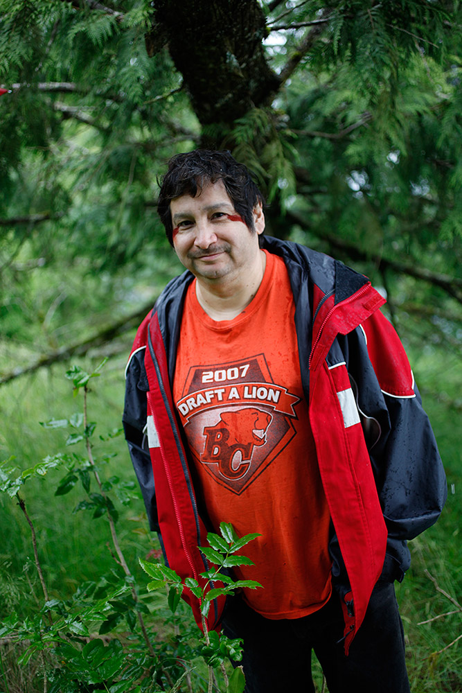 A man stands in front of a tree in a forested area; he has red ochre on the outer corners of each eye.