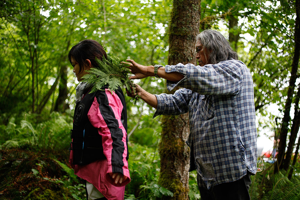 A man holds a cedar branch in his hands, and brushes it against a woman’s back.