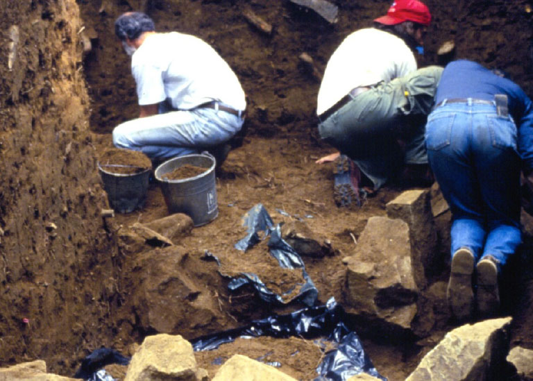 Several people are working inside an excavated archaeological mound. They are surrounded by dirt walls, rocks, and buckets filled with dirt.