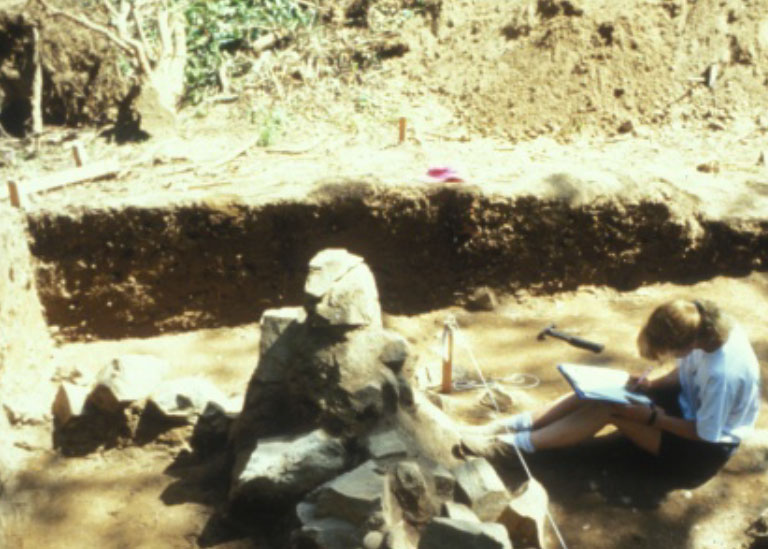 A woman takes field notes in front of a rock formation. She is seated beside an excavated trench.