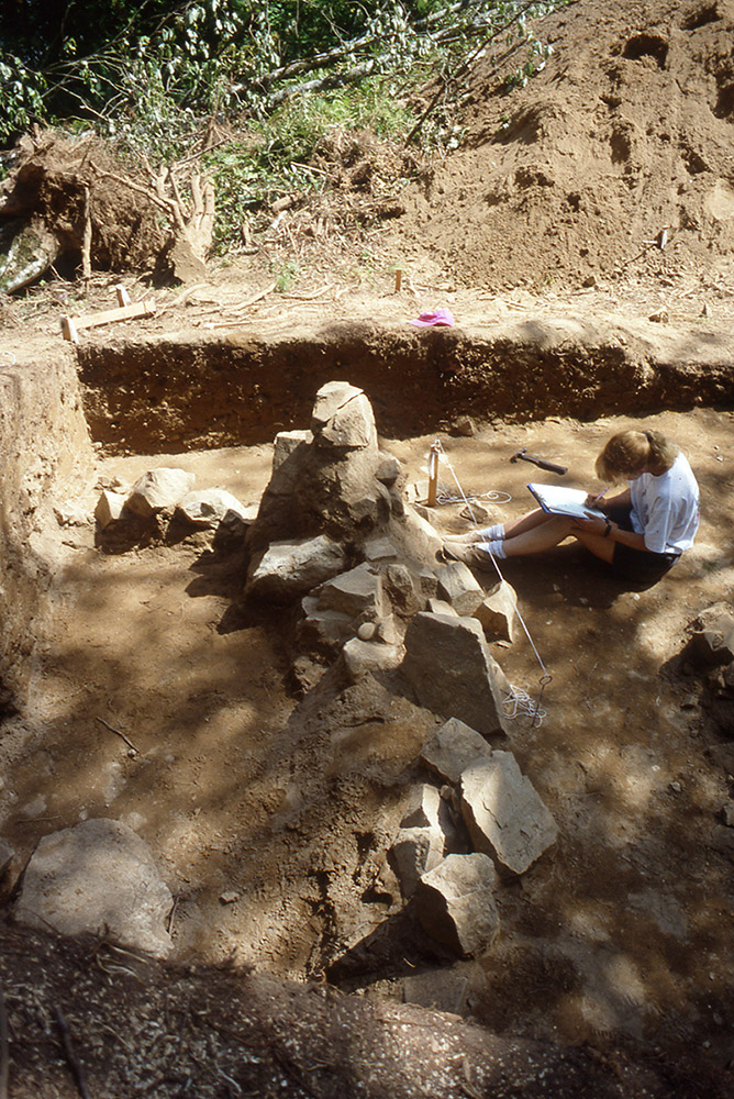 Une femme rédige des observations de terrain devant une formation rocheuse. Elle est assise à côté d’une tranchée.