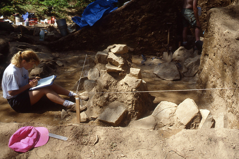 A woman takes field notes in front of a rock formation. She is seated in front of an excavated mound. 