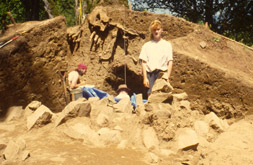 Plusieurs personnes sont réunies près d'un vaste tas de terre où ont lieu des fouilles. Le tertre mesure trois mètres de haut et a été divisé en deux dans sa longueur. Il y a plusieurs grosses roches près de la base du tertre.