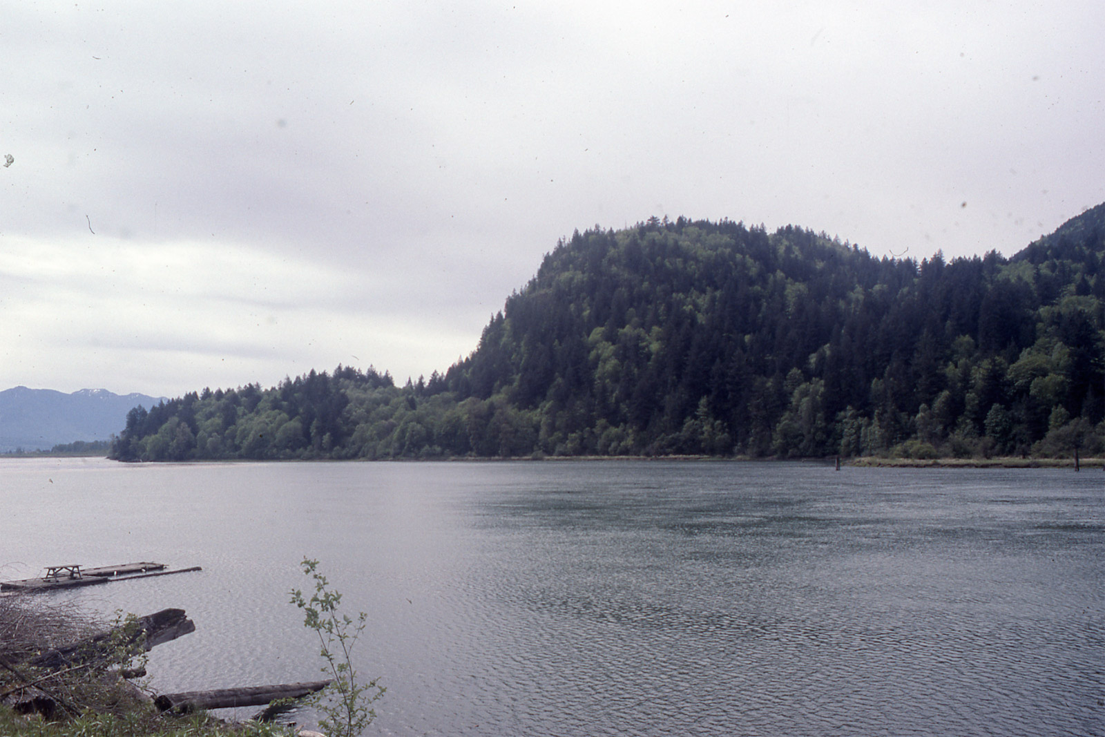 A grey river in the foreground with a hill in profile across the water, low and then rising to a hump
