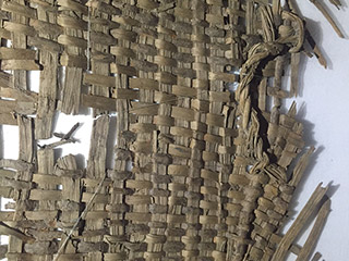 A fragment of woven basketry. The weaving material is light greyish brown.