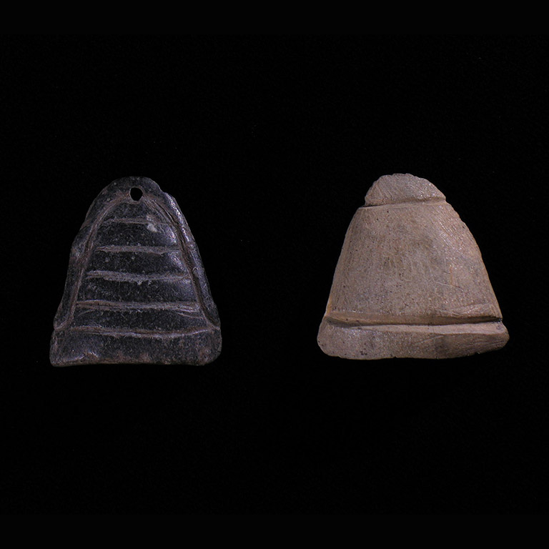 Deux petits morceaux de pierre sculptés dans une forme triangulaire. Le contour de la pierre noire est gravé et comporte cinq lignes gravées à l’horizontale; la partie supérieure est percée. La pierre pâle a des lignes gravées près du haut et du bas de la pierre.