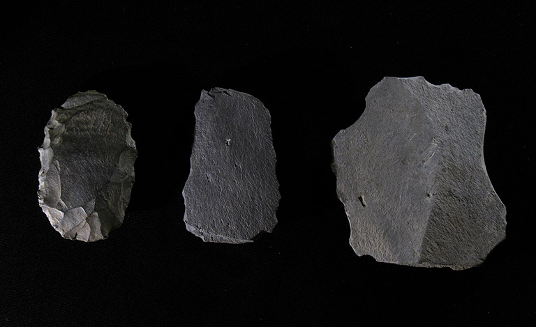 Trois morceaux de pierre gris foncé avec des bords aiguisés, posés sur un fond noir.