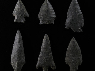 Six pointes de flèche de grosseurs et largeurs différentes faites de pierre foncée, posées sur un fond blanc.