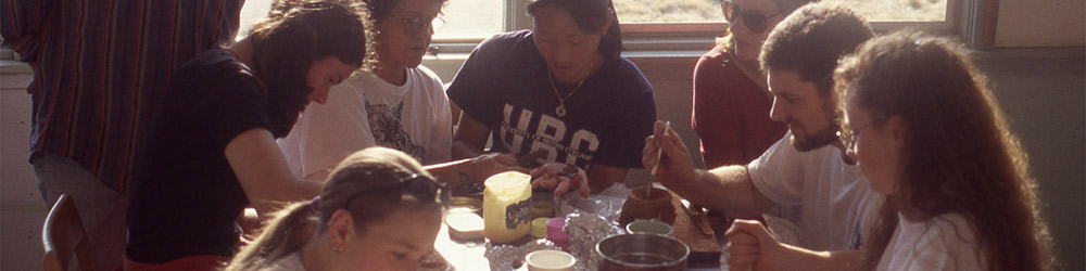 Many students are gathered at a rectangular table, using various tools and materials, including red ochre.