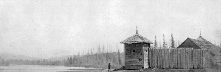 Au milieu de la photo, la structure d’un fort avec un mur de billots devant et une personne qui se tient à l’extérieur du fort. Il y a des conifères le long de la rive au loin.