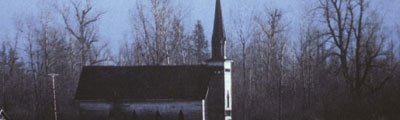 A side view of a small church in a field. In the background is a forest and a snow-covered mountain.