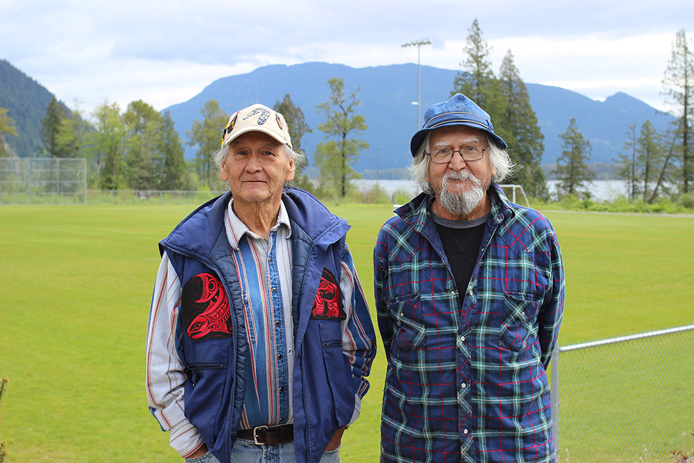 Two men stand in front of a soccer field. The man on the left is wearing a blue vest with red designs on it. The man on the right has his hands behind his back and is wearing a blue plaid shirt.