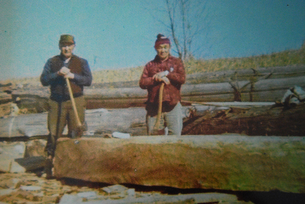 Two men stand behind a log. Both men are holding axes that are resting on the log.